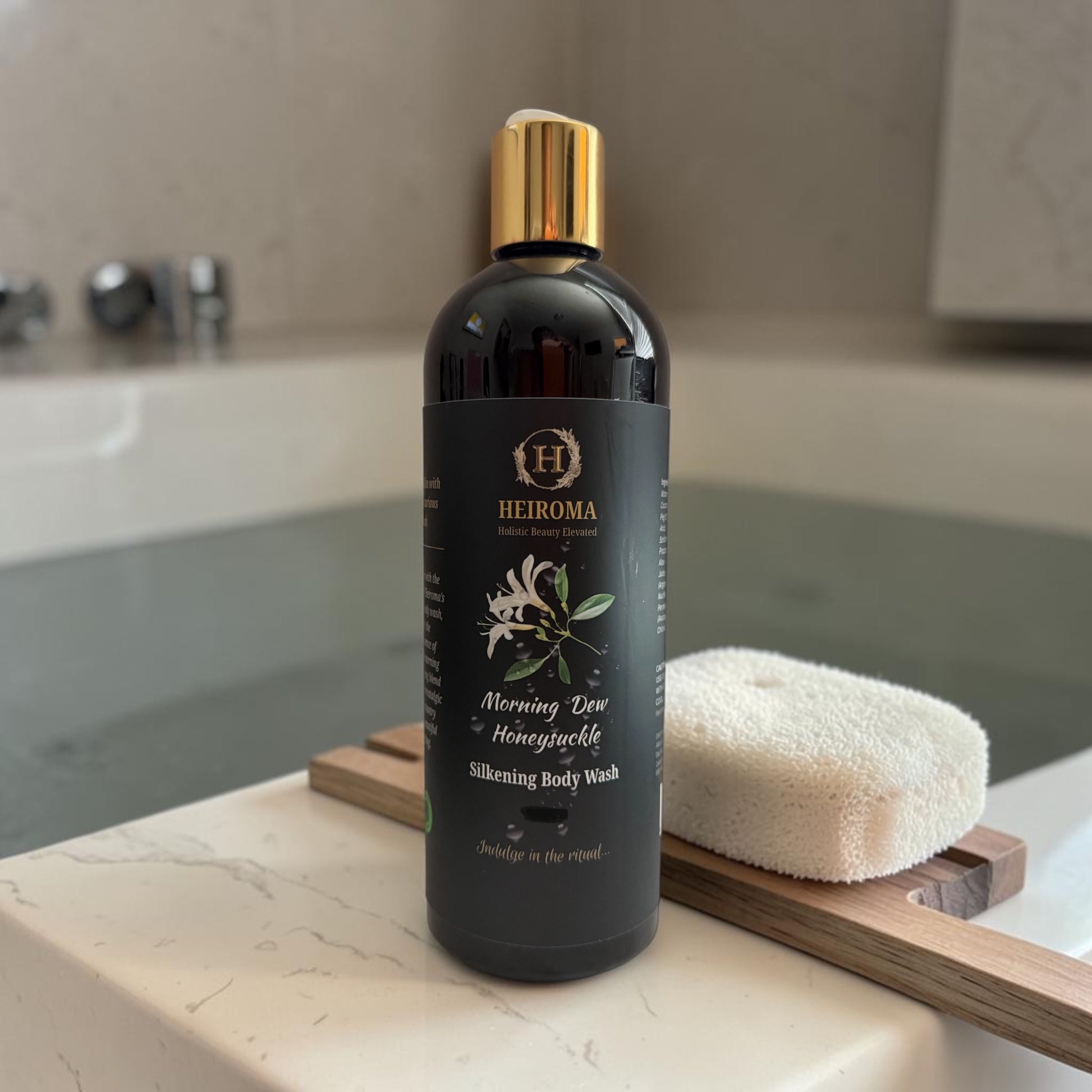 A bottle of HEIROMA Morning Dew & Honeysuckle Silkening Body Wash stands on a marble bathroom counter next to a white sponge on a wooden stand.